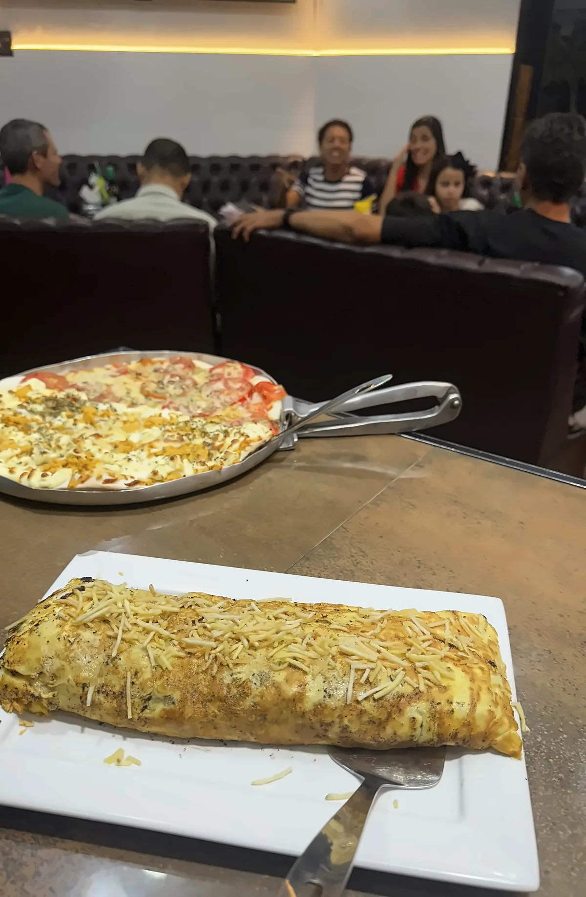 Mesa no salão da Lussan à noite, com pizza servida e o salão ocupado ao fundo.