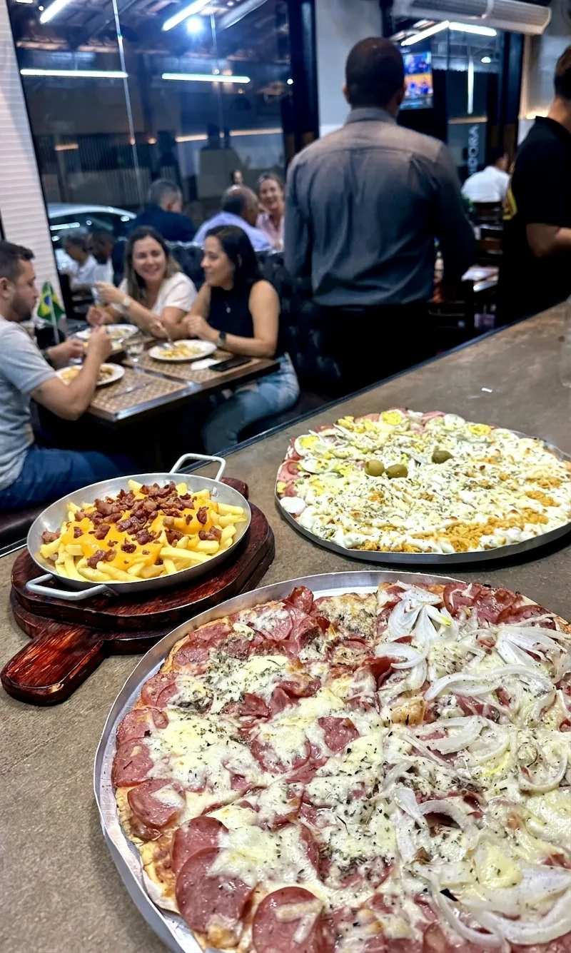 Balcão da Lussan à noite: duas pizzas em bandeja e uma porção de fritas, com o salão ao fundo.