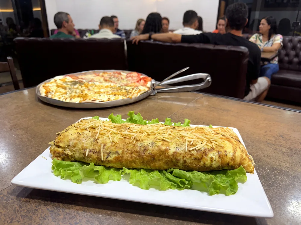 Mesa da Lussan com pizza na bandeja e omelete recheado na travessa, e o salão ocupado ao fundo.